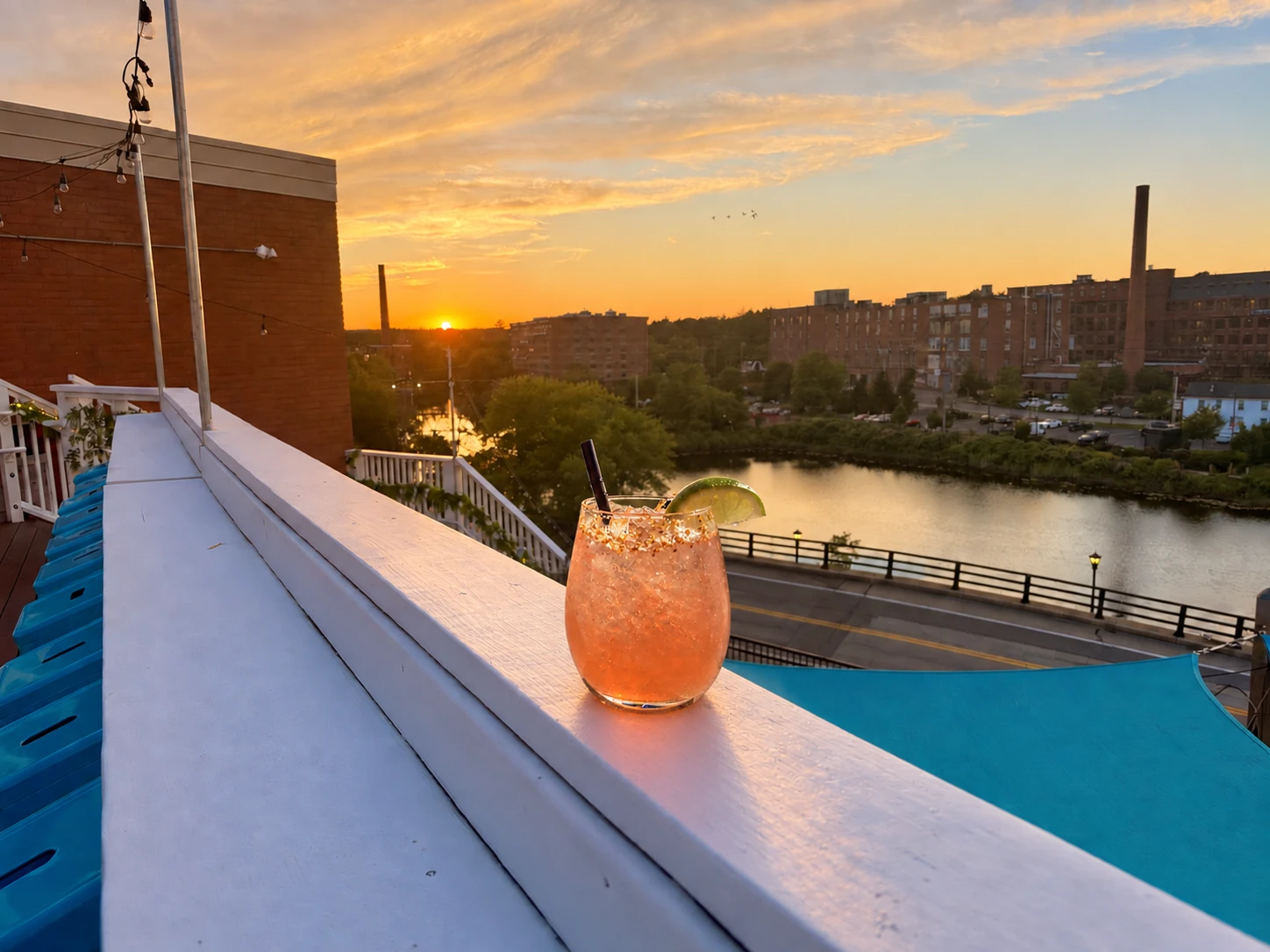 A cocktail resting on a rooftop railing at sunset, looking out over the river and the mill buildings
